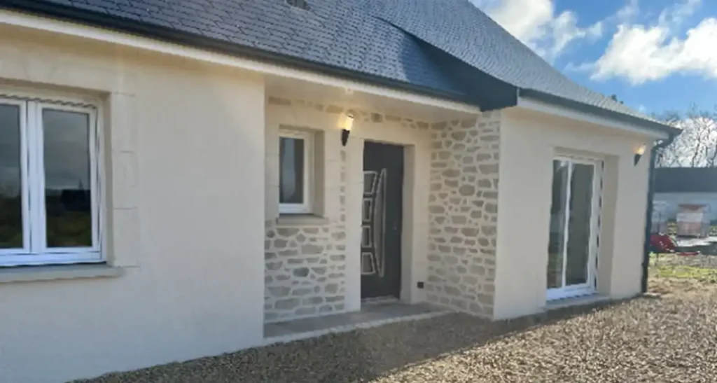 Maison de plain-pied avec une façade extérieure en pierre claire et enduit crème, un toit sombre, une porte d'entrée moderne, deux fenêtres et une cour en gravier. Le ciel est partiellement nuageux à l'arrière-plan.