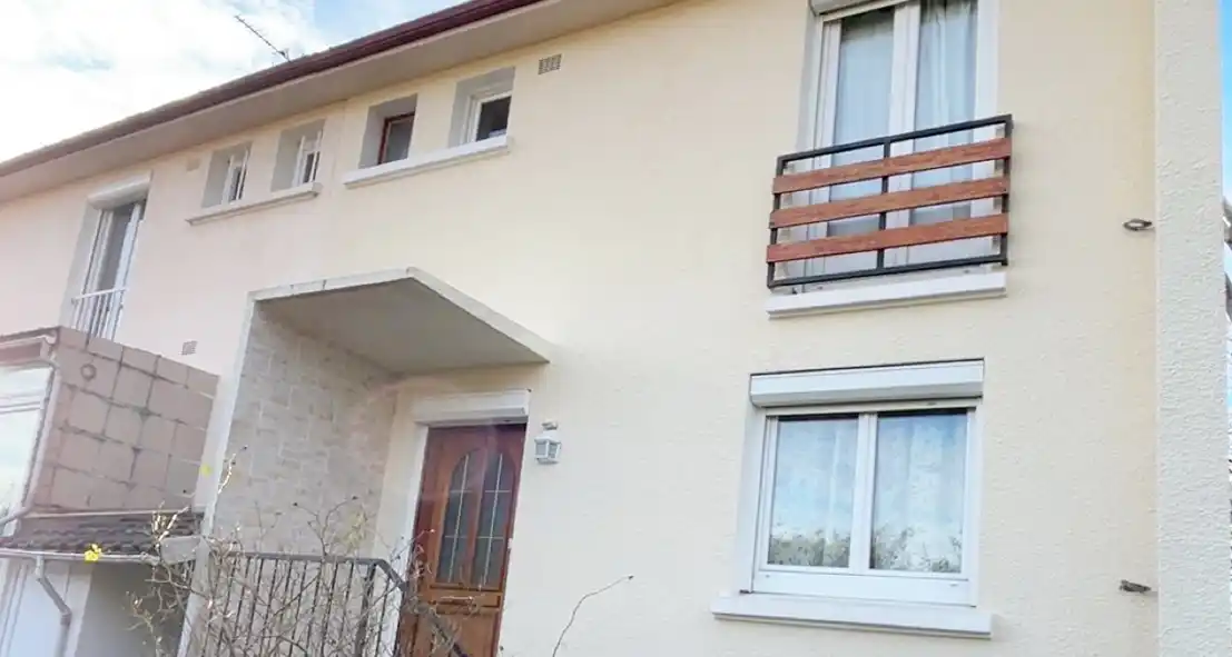 Rénovation de façade sur maison à étage – Girault Ravalement Une maison de deux étages de couleur crème avec une porte d'entrée en bois, de grandes fenêtres et un petit balcon avec des lattes en bois, avec une entrée couverte. L'extérieur simple présente des bordures blanches autour des fenêtres, magnifiquement mises en valeur par un récent ravalement de façade.